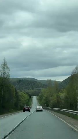 The video shows a car driving on a two-lane road. The road is bordered by green trees on either side, with hills in the background, under a cloudy sky. Two cars are ahead of the camera car, a red car and a white car. A guardrail runs along the right side of the road.
