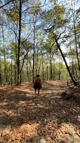 The video shows a woman walking along a wooded path covered in fallen leaves. She is wearing a black sports bra and black shorts. The path is surrounded by tall trees, some of which are leaning. The sky is visible through the branches. The camera follows the woman from behind, then she briefly turns the camera towards herself.
