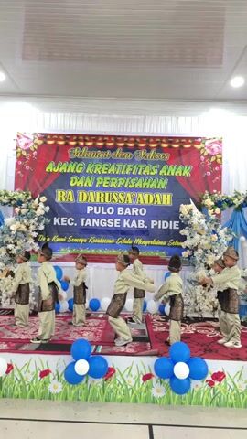 A group of young boys in traditional clothing are performing a dance on a stage. The stage is decorated with a banner that reads "SELAMAT dan SUKSES AJANG KREATIFITAS ANAK DAN PERPISAHAN RA DARUSSA'ADAH PULO BARO KEC. TANGSE KAB. PIDIE". There are also balloons and floral arrangements on the stage. The boys are all wearing similar outfits, consisting of a light-colored tunic, pants, and a dark sash. Some of them are wearing hats. They are in various poses, with some of them reaching out their arms. The audience is not visible in the frame. The lighting appears to be artificial, possibly from stage lights. The background is a white wall with some dark ceiling elements visible at the top. The overall atmosphere seems festive, likely an event or performance at a school or community center.