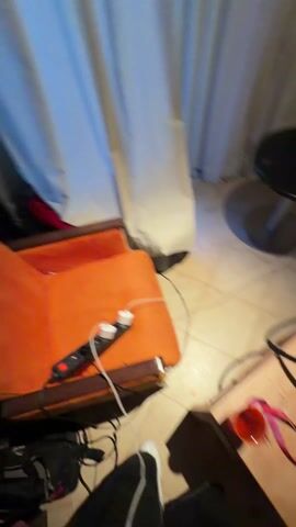 The video shows a young man sitting on a beige couch in a hotel room. He is shirtless, wearing shorts and sandals. He is using a small electronic device with colorful buttons and screens. A table next to the couch holds several disposable cups and other items. There is a hookah on a stand nearby. A framed soccer jersey hangs on the wall behind the couch. The room has white walls and beige curtains.
