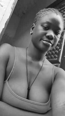 A close-up, low-angle, black and white video shows a young Black woman with short, braided hair. She is wearing a thin-strapped tank top and a necklace with a dark pendant. The woman opens her eyes and glances directly at the camera with a slight smile. The background includes a textured door and part of a wall.