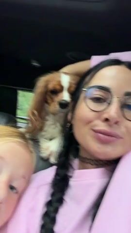 In this close-up, selfie-style video, a woman with black hair in two braids, wearing glasses and a pink sweatshirt, is in the foreground. Next to her, to her left, a young girl with blonde hair rests her head on the woman's shoulder, her eyes closed. Behind them, perched on the seat, is a Cavalier King Charles Spaniel with brown and white markings. Further in the background, another person, likely a child, is partially visible. The scene appears to be inside a vehicle, possibly a car, judging by the headrests and the interior lighting. The background outside the vehicle suggests a daytime setting, with greenery visible, hinting at a rural or suburban environment. The overall mood seems relaxed and intimate, with the girl's closed eyes suggesting she might be sleeping or resting comfortably. The woman is looking towards the camera with a slight smile. The video is captured during what appears to be daylight hours. No text is visible in the video.
