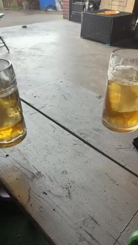 The video shows two large beer mugs filled with golden lager and topped with frothy white heads. The mugs are held by unseen individuals and are positioned on a rustic wooden table. The lighting suggests an outdoor or semi-outdoor setting, possibly a patio or garage, with concrete flooring visible in the background and some outdoor furniture. The camera appears to be looking down at an angle, focusing on the beer mugs. The overall impression is one of a casual, possibly social, moment.