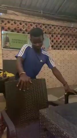 A young man dances behind stacked chairs at an outdoor bar, smiling and gesturing to the music. He wears a blue Adidas t-shirt and colorful shorts. Posters advertising beer are visible on the wall behind him, along with decorative latticework. The man’s hands are adorned with bracelets.