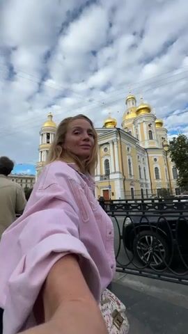 A blonde woman in a pink jacket is taking a selfie in front of a yellow church with golden domes. The church is on a street with cars driving by. There is a black metal fence in the foreground. The sky is cloudy.