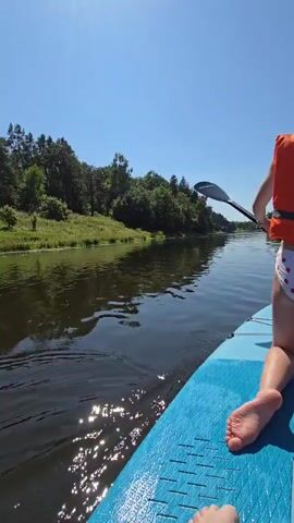 A person on a paddleboard is shown from the knees down. The person is wearing a life vest and is paddling in a river. The river is surrounded by trees and grass. The sky is blue and clear.