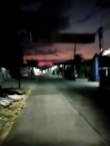 This video appears to be a first-person perspective of someone riding a motorbike through a street in Menjing, Indonesia, at dusk. The sky is a deep purple and pink, indicating the end of daylight, and dark clouds are visible. Streetlights and lights from buildings illuminate the road, which is paved and lined with houses, shops, and trees. 

Several motorbikes are seen on the road, and one person on a bicycle is also visible. The overall mood is calm and serene, characteristic of a quiet evening in a residential area. The location cues, such as the style of houses and the presence of palm trees, along with the mention of Menjing, Indonesia, help establish the setting. There is no visible text in the video.