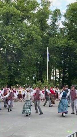 The video shows a group of people in traditional clothing dancing in a circular formation outdoors. The dancers are dressed in folk costumes, with women wearing long skirts and blouses, and men wearing vests and trousers. They are holding hands and moving in a synchronized manner. In the background, there are tall trees and a flagpole with a flag. There is also a stage with white streamers hanging from it. The lighting suggests it is daytime, possibly late afternoon or early evening. The atmosphere appears festive.