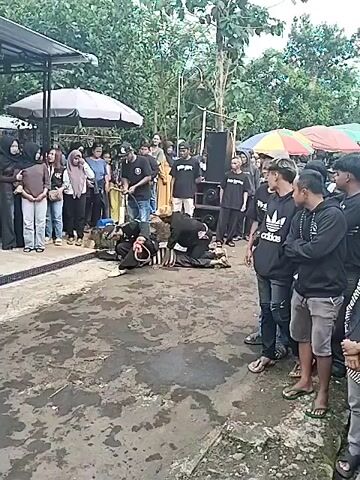 The video depicts an outdoor scene with a group of people gathered around what appears to be a performance or ritual.  The ground is concrete and appears wet in places.  Most people are watching a figure lying on the ground, wearing a striped shirt and dark pants.  Two other individuals, dressed in black, are kneeling beside the person on the ground, seemingly attending to them or participating in the ritual.  The crowd consists of both men and women, some wearing casual clothes like jeans and t-shirts, while others are dressed in traditional attire.  Several people are holding umbrellas for shade.  In the background, there is a speaker, various tables and umbrellas, and lush greenery.  Some people are smiling or have neutral expressions, suggesting a mix of curiosity and engagement with the event.