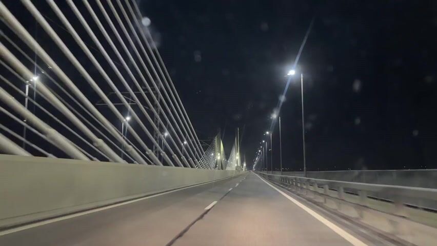 A car drives on a bridge at night. The bridge has cables and lampposts on both sides. There are white lines on the road dividing the lanes. It appears the camera is situated inside the vehicle, looking outwards from the windshield.  The road is mostly empty. The sky is dark and clear. There may be moisture on the windshield.