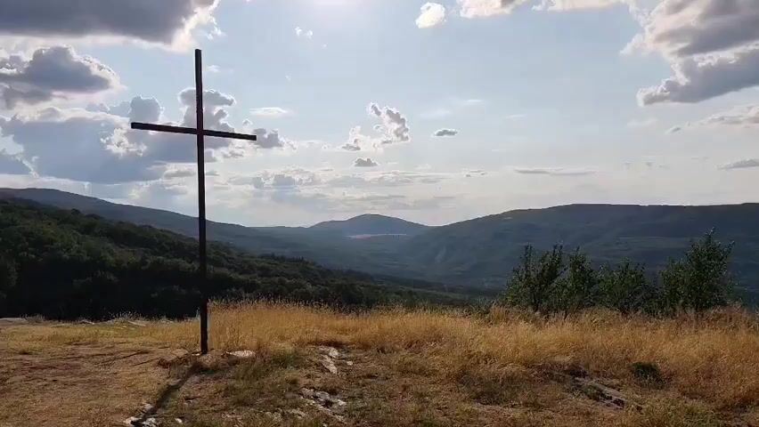 This is a video that pans across a landscape with a cross in the foreground. The cross is made of dark metal and stands on a hill. Behind the cross, there are rolling hills covered in trees, and in the distance, there are more hills and mountains under a partly cloudy sky. The sun is visible in the sky, casting a bright light. The ground in the foreground is covered in dry grass and rocks. The camera moves from left to right, showing different angles of the cross and the surrounding scenery.