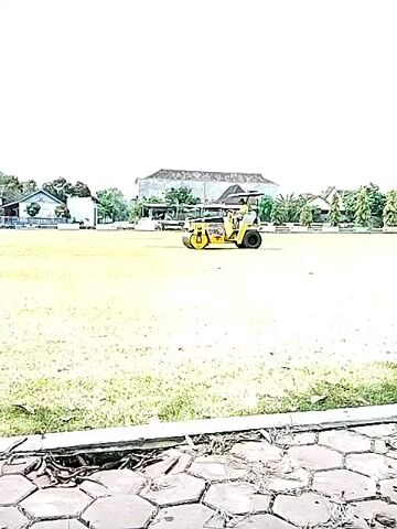 The video shows a yellow roller moving across a green field.  The camera slowly zooms in.  The field has buildings and trees in the background. The sky is blue and has white clouds.  There is a black bird flying in the sky. In the foreground are grey hexagon-shaped pavers. A drain is visible in the pavers.
