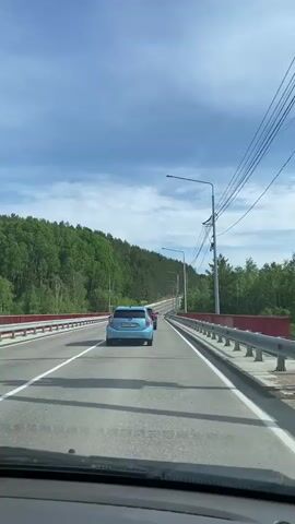 The video shows a view from inside a car driving on a paved road with white lane markings. The road is bordered by metal guardrails. On the left side of the road, there is a dense forest with green trees covering a hillside. On the right side, there are also trees and utility poles with wires running along them. Several cars are visible on the road. A light blue car is in front of the camera's car, and a darker red car is ahead of the blue car. The sky is blue with some white clouds. The overall scene depicts a drive through a wooded area on a sunny day.