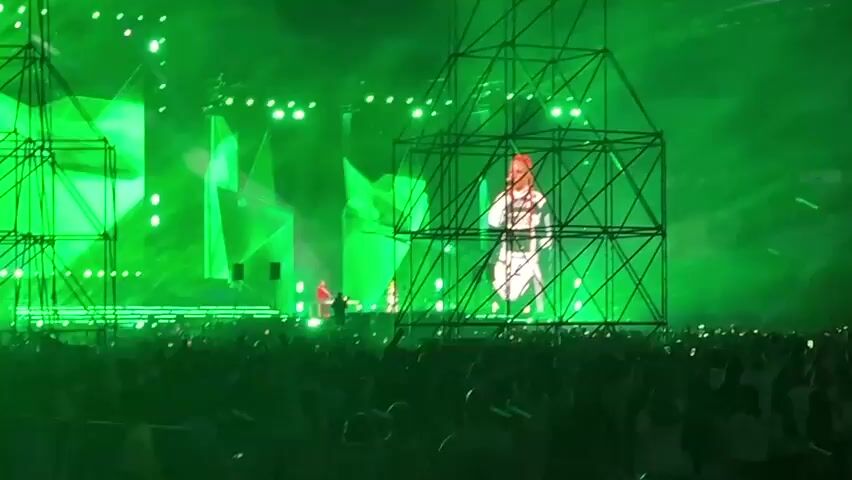 A concert stage bathed in green light features a large screen displaying a stylized image of a person wearing a white outfit with black stripes. In front of the screen, two figures in red clothing stand on the stage. To the left and right of the stage are tall, black metal scaffolding structures. The foreground is filled with the silhouettes of a large audience, many holding up phones, all illuminated by the green stage lighting and light-emitting devices from their phones. The overall atmosphere is that of a live musical performance with a significant crowd.
