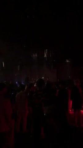 The video shows a crowd of people in a dark, dimly lit environment, likely a nightclub or concert venue. The lighting consists of spotlights and ambient colored lights, creating a moody atmosphere. Several people are visible, appearing to be dancing or socializing. There are speakers and lighting equipment suspended from the ceiling. The overall impression is that of a lively event with a significant gathering of individuals.