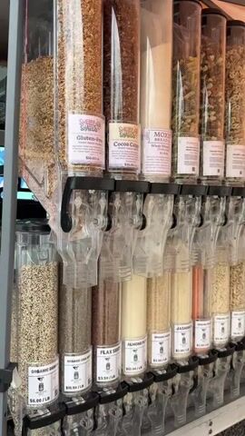 The camera slowly pans across a bulk food dispenser at what appears to be an outdoor market or store.  The dispenser features clear plastic containers filled with a variety of grains, flours, pasta, cereals, nuts, dried fruit, and candy.  Each container is labeled with the product name and price per pound. The containers are arranged in rows and tiers, with a dispensing mechanism at the bottom of each.  The bottom tier is made of clear plastic, while the middle and top containers are covered with the product label. A sign at the end invites viewers to follow the store on social media.