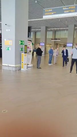 The video shows the interior of a modern, well-lit building, likely an airport or train station. The floor is tiled with a marble-like pattern. The walls and ceiling are white and industrial, with exposed lighting fixtures and ventilation systems. Several pillars, covered in grey panels, are visible.

In the background, there are signs with text and icons, suggesting directional information or advertisements. One sign displays a yellow QR code. Another shows a green directional arrow and an accessibility icon. There are also some shops or kiosks with merchandise displayed behind glass windows.

Several people are present. In the mid-ground, a man in a dark jacket and grey pants is standing near a kiosk, looking at his phone. To his right, three older men in casual attire are standing in a line, appearing to wait for something. Further to the right, a man in a dark suit is walking and speaking on his phone, carrying a black bag.

In the foreground, a green arrow is visible on the floor, possibly indicating a pathway or social distancing marker. The overall impression is one of a clean, functional, and busy public space.