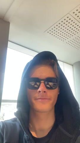A young man wearing a dark blue hooded sweatshirt and black sunglasses stands facing the camera. The hood is up and pulled forward on his head. The background shows a white, patterned ceiling with a ventilation grill, and a brightly lit window to the left. The man remains still and looks directly at the camera throughout the video.