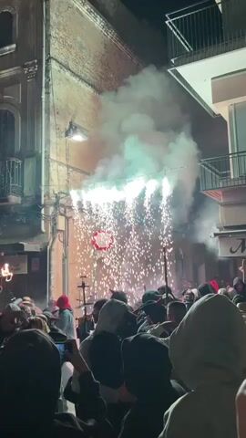 The video captures a nighttime outdoor event in a narrow, urban street lined with multi-story buildings. The scene is dominated by a dramatic pyrotechnic display erupting from a structure or platform across the street. White and orange sparks rain down, accompanied by a significant amount of white smoke that billows upwards. A red circular object is visible within the sparks. A crowd of people, densely packed and facing the display, fills the street. Many are wearing hooded sweatshirts, and some appear to be holding up phones to record the event. A person on the left side of the frame holds a lit torch. The atmosphere suggests a festive or celebratory occasion, possibly a festival or parade, with a strong element of fire spectacle.