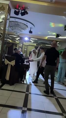 A room filled with people dancing. There are several tables with seated individuals, and a group of people dancing in the center. Some are standing, some are dancing, and some are eating. The room has checkered floor tiles. The ceiling features modern lighting, including a chandelier. Some of the people are wearing formal wear. There are sound and light systems present.