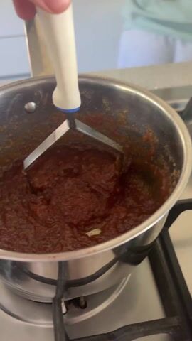 A hand is using a metal potato masher to mash a dark brown mixture in a stainless-steel pot on a black gas stovetop. The camera angle is slightly overhead. The stovetop is clean, and the mixture appears to be simmering. The mashed mixture looks thick and concentrated. The camera rotates slightly to show items reflected in the bottom of the pot, including a whisk, a pink container, and a pink tray with small white candies. The background shows a white kitchen counter with a lemon motif and a person in a light blue shirt visible in the distance.