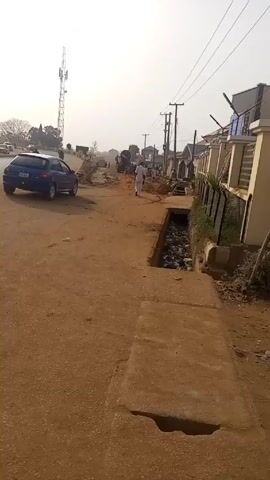 The video shows a dusty, unpaved road in a residential area, likely in a developing country. A blue car is parked on the left side of the road, and several people are walking in the distance towards what appears to be a construction site. On the right, there are houses with fences and power lines overhead. A concrete drainage ditch runs along the side of the road, filled with debris. A motorcycle carrying three people passes by on the road. The sky is overcast, creating a hazy light.