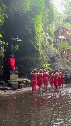 A group of people in red sarongs stand in a shallow stream in front of a moss-covered rock face. There is a stone altar draped with red fabric and a decorative umbrella. A statue of a figure is partially visible behind the altar. Further up the rock face, another statue and a small temple structure are visible. The environment is lush and green with vegetation growing on the rocks. The people are facing the altar and appear to be participating in a ritual or ceremony. The water is clear and reflects the surrounding greenery.