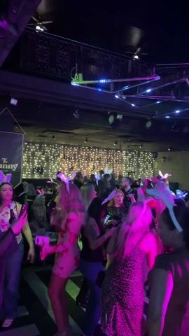 The video shows a lively scene inside a nightclub, bustling with activity. Many attendees, predominantly women, are wearing glowing bunny ears. They are dancing and interacting with each other, illuminated by vibrant pink and blue lighting. The club has a mezzanine level, elaborate lighting fixtures and a backdrop of twinkling fairy lights. Banners for "The Bunny Club" are visible. Some people are recording the event on their phones. The environment is loud and energetic.