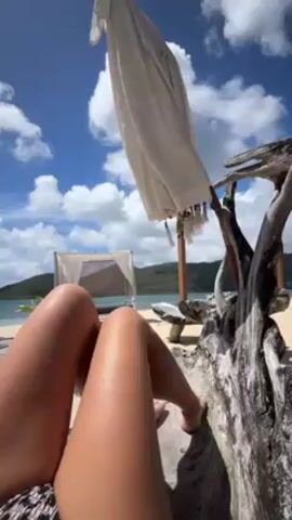 The video shows a first-person view of a person lying down, presumably on a beach. The lower legs are visible in the foreground, appearing tan. Behind the legs, the scene opens up to reveal a tropical beach. There is a white canopy bed with sheer curtains on the left side of the frame, set up on the sand. In the background, there is an island or landmass covered in lush greenery. The sky is blue with scattered white clouds, suggesting a sunny day. A white umbrella or parasol hangs overhead, slightly obscuring the view. To the right of the legs, there is a piece of driftwood. The camera remains still throughout the video.