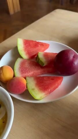 The video begins with a close-up shot of a white plate filled with sliced watermelon, two small peaches or apricots, and a dark plum. Next to the plate is a white bowl containing a fruit salad with what appears to be yogurt, orange slices, kiwi, and shredded coconut. The camera then pans slightly to the left, revealing more of the fruit salad. The scene then transitions to a wider shot of a restaurant interior. Wooden tables and chairs are visible, with some diners seated at them. Light-colored curtains are hung between sections of the restaurant, and wooden beams are present in the ceiling. The flooring is made of dark wood. The final shots offer a broader view of the restaurant, showing more tables, chairs, and patrons, as well as a bar or serving area in the background. The overall impression is of a casual dining establishment.