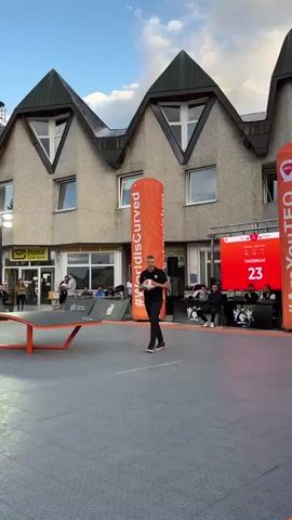 A man in a black t-shirt and pants stands in the center of an outdoor paved area, holding a ball. Behind him, a building with a gray facade and peaked roofs stands under a blue sky with scattered clouds. Two tall, inflatable orange pillars with the text "#WorldIsCurved" and "#AreYouTEQ" flank a large red digital screen displaying a countdown timer that reads 22, then 21, 20, and 19, decreasing by one each second. Several people are seated behind the screen. To the left, a table tennis-like apparatus with an orange surface is visible. People are scattered around the area, some standing and others seated. The overall environment appears to be an outdoor event space.