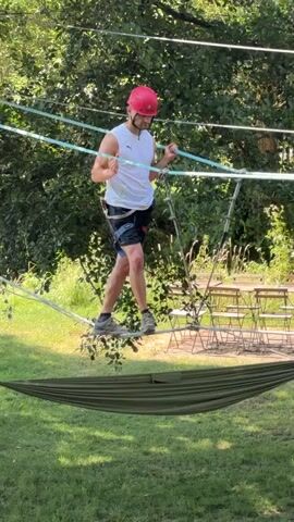 A man wearing a pink helmet, white tank top, and black shorts is walking on a rope bridge suspended between trees. He is holding onto ropes on either side for balance. Below the rope bridge, a green hammock is stretched out. The man is in the middle of crossing the bridge. In the background, there are more trees and some outdoor seating with tables and chairs. The lighting suggests it is daytime and sunny.