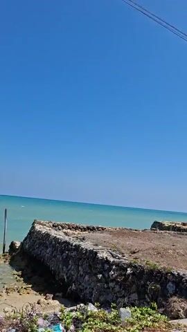 The video shows a coastal landscape under a clear blue sky. The foreground is a dry, sparsely vegetated area with debris. In the middle ground, the turquoise ocean meets the shore. Along the coast, there are remnants of structures, including concrete foundations and what appears to be a partially collapsed building with a corrugated metal roof. Further down the coast, there are several small, simple buildings with white walls and tiled roofs. Overhead, power lines are visible against the sky. The camera pans slightly to the right, revealing more of the coastal area and the scattered structures.