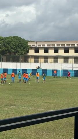 A group of male soccer players wearing orange and blue jerseys are on a green field. They are spread out and passing a soccer ball around. A large green inflatable ball with text is visible in the background, near a white goal with a net. A light brown building is behind the goal, and trees are on the left. The sky is cloudy.