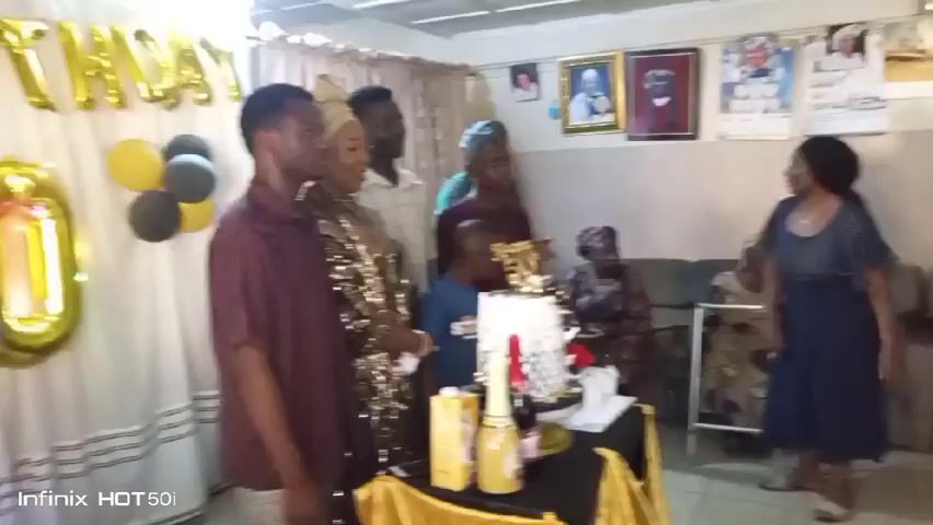The video shows a small indoor gathering for a 50th birthday celebration. The backdrop is decorated with a white curtain, gold "HAPPY BIRTHDAY" balloons, gold "50" balloons, and black and gold balloons. Several people are present, including a woman wearing a gold dress and head wrap. There is a birthday cake on a table, surrounded by champagne bottles. People are standing around, seemingly getting ready to celebrate. An "Infinix HOT 50i" watermark is visible in the bottom left corner of the video.