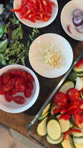 The media shows a top-down view of various chopped vegetables and herbs arranged on a wooden surface. In a large glass baking dish, sliced zucchini, cherry tomatoes, red bell pepper strips, and red onion wedges are prepared. To the left, a white bowl contains halved cherry tomatoes, and a small white plate holds minced garlic. Another small white plate displays sliced red onion. A bunch of fresh basil and oregano sprigs are also visible. In the background, there are jars and containers, possibly spices or pantry items. The scene appears to be in a kitchen, likely for food preparation. The lighting suggests it could be daytime, with natural light. No people are directly visible, but a person's foot is slightly in frame at the bottom right, suggesting someone is preparing the meal. There is no discernable text in the media.