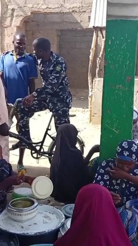 Here is a description of the video:

The video shows an outdoor scene with several people gathered in what appears to be a public or semi-public space. In the foreground, a group of women are sitting around a large tray or low table filled with bowls and food. They are eating. Some are wearing hijabs or head coverings. A man in a blue shirt is standing near a bicycle. A man in camouflage patterned clothing is sitting on the bicycle. He is conversing with the man in the blue shirt. In the background, there are other people walking around. Motorbikes are parked under a corrugated metal awning. A woman in a long red robe is carrying a tray of greens on her head. Several children are nearby. There is a brick wall and green pillar in the background. The atmosphere appears relaxed.