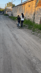 Child cycles dirt road with adult companion in Gyumri
