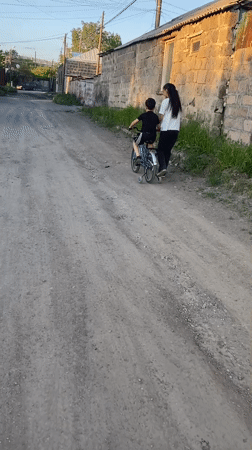 Child cycles dirt road with adult companion in Gyumri