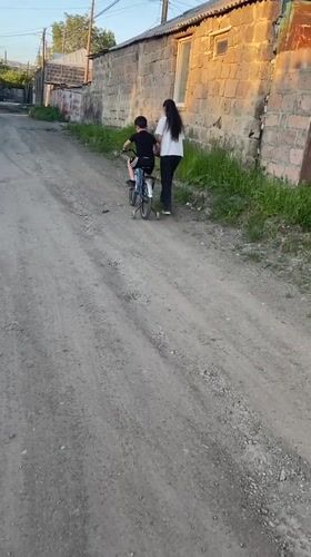 Child cycles dirt road with adult companion in Gyumri