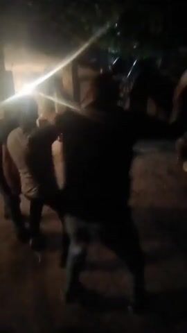 The video shows a group of people engaged in a lively dance. The scene is set outdoors at night, indicated by the darkness and artificial lighting. The lighting, which appears to be from streetlights or lamps, casts long shadows and creates a somewhat hazy atmosphere. Several individuals are actively dancing, some in pairs and others in a more individualistic style. Their movements are energetic, suggesting a festive or celebratory mood. In the background, more people are visible, some appearing to be observing or participating in the event. There are also objects that could be furniture or equipment, and hints of structures suggesting a built environment, possibly a courtyard or an outdoor gathering space. The overall impression is of a social gathering where music and dancing are taking place, possibly a party or a community event. The time of day is clearly night. The weather is not discernible. There is no visible text in the video. The location context indicates Aragatsavan, Armenia.