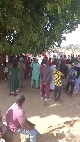 The video shows a gathering of people under the shade of a large tree. The group appears diverse in terms of clothing, with some wearing traditional garments and others in more modern attire. A man in a light blue outfit plays a drum slung across his shoulder. Some people are dancing and moving to the rhythm of the music. The ground is dusty, and the background shows hints of a rural setting with more people and structures further away. One man is seated in the foreground, watching the scene.