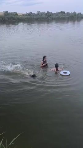 In this media file, three individuals are present in a body of water. Two appear to be young women, one in a black swimsuit and the other in a patterned swimsuit. The third individual is partially submerged, with only their head and shoulders visible, and appears to be wearing a dark swimsuit. A white and blue inner tube is also visible in the water. The scene takes place on a body of water, likely a river or lake, with trees lining the far bank. The sky is overcast, suggesting a cloudy or partly cloudy day. The water is calm with some ripples, indicating gentle movement. The overall mood appears to be relaxed and leisurely, with the individuals enjoying a swim. The location is identified as Ciumai, Moldova, providing geographical context to the scene. No text is visible within the media file.