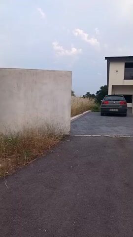 The video shows a static shot of a driveway with two cars parked in front of a modern house. The driveway is paved with dark interlocking stones. To the left of the driveway is a white concrete wall with overgrown grass and weeds. To the right of the driveway is a two-story modern house with a white facade and dark-framed windows. The house is partially covered in green ivy and has a flat roof. One car, a dark-colored hatchback, is parked directly in front of the house. Another silver sedan is parked to the right of the hatchback. The sky above is overcast with some clouds. There are no people visible in the video.