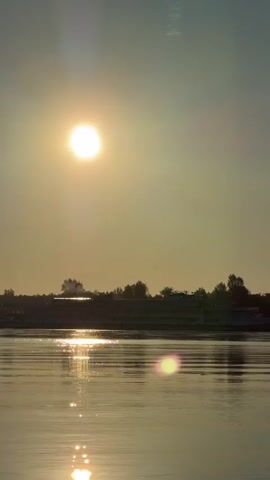 The video shows a wide shot of a body of water during sunset. The sun is a bright, diffused orb in the hazy sky, casting a golden glow and reflecting on the rippling surface of the water. In the middle ground, a long, multi-decked cruise ship is visible docked or moving slowly along the far bank. The shoreline behind the ship is lined with trees and vegetation. The overall atmosphere is calm and serene.
