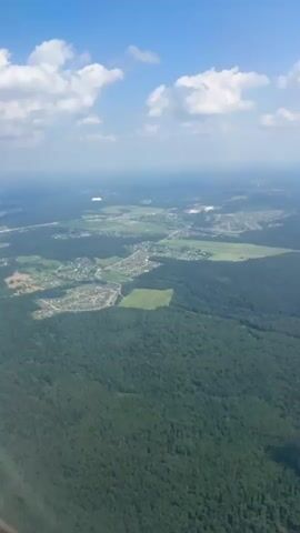 This is an aerial view of a rural landscape under a bright blue sky dotted with fluffy white clouds. The scene depicts a vast expanse of dense green forest interspersed with small clearings and scattered settlements. In the foreground, the dark green canopy of the forest dominates, suggesting a temperate climate.

Further into the distance, the landscape opens up to reveal a mosaic of agricultural fields, some appearing cultivated and others fallow, alongside clusters of houses that form villages. A winding river or road is faintly visible cutting through the terrain. The overall impression is one of a peaceful, sprawling countryside.

The time of day appears to be daytime, likely late morning or early afternoon, given the bright sunlight casting shadows and illuminating the scene. The weather seems clear and fair, ideal for aerial observation. The location is identified as Borduki, Russia, which aligns with the visual cues of a predominantly forested and agricultural area. No people or specific objects are distinctly identifiable from this altitude, but the presence of human settlements and agricultural activity indicates human presence and land use. The image evokes a sense of tranquility and the vastness of nature. No visible text is present in the media.