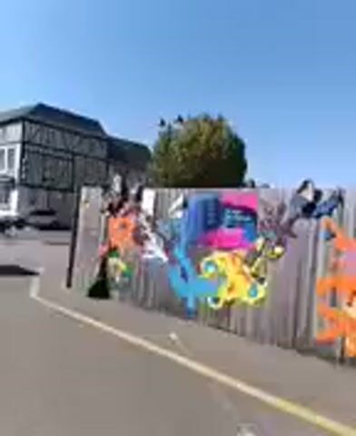 The video pans across a wooden fence decorated with vibrant, colorful graffiti. The graffiti features abstract shapes and cartoonish figures in bright blues, pinks, yellows, oranges, and purples. Behind the fence, a two-story building with a dark-tiled roof and white-framed windows is visible. The sky is clear and blue. To the left of the building, another building with a similar architectural style can be seen. The ground in front of the fence is paved with asphalt and has a yellow line marking. The camera movement is steady, suggesting it is being held or mounted on a moving object like a vehicle.