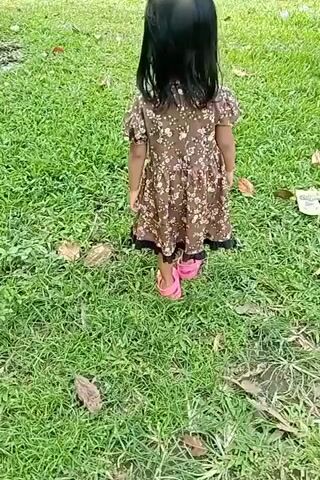 A young girl with black hair, wearing a brown floral dress and pink sandals, walks across a grassy area. She is seen from behind and from a side angle as she moves. The environment appears to be an outdoor park or lawn, with green grass and some fallen leaves visible. In the background, there are some light-colored fabric objects, possibly tents or blankets, and a tree trunk. The lighting suggests it is daytime.