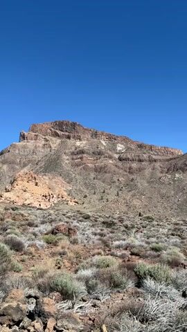 The video pans across a barren, arid landscape dominated by a large, layered mountain under a clear blue sky. The foreground is covered in dry, scrubby vegetation and rocks. The mountain features distinct geological strata, indicating erosion and different rock types. The overall impression is one of a remote, undeveloped, and dry environment.