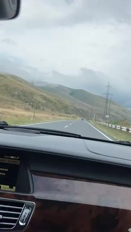 This video provides a first-person perspective from inside a car driving on a road. The road is asphalt with white lane markings. On the right side of the road, there is a guardrail and several tall power line towers. The landscape features rolling hills with sparse vegetation. The sky is overcast with clouds, and there are some lights or reflections visible on the windshield. The interior of the car shows a dashboard with a navigation screen displaying some text, wood trim, and a window pillar. The car appears to be in motion, as indicated by the passing scenery.